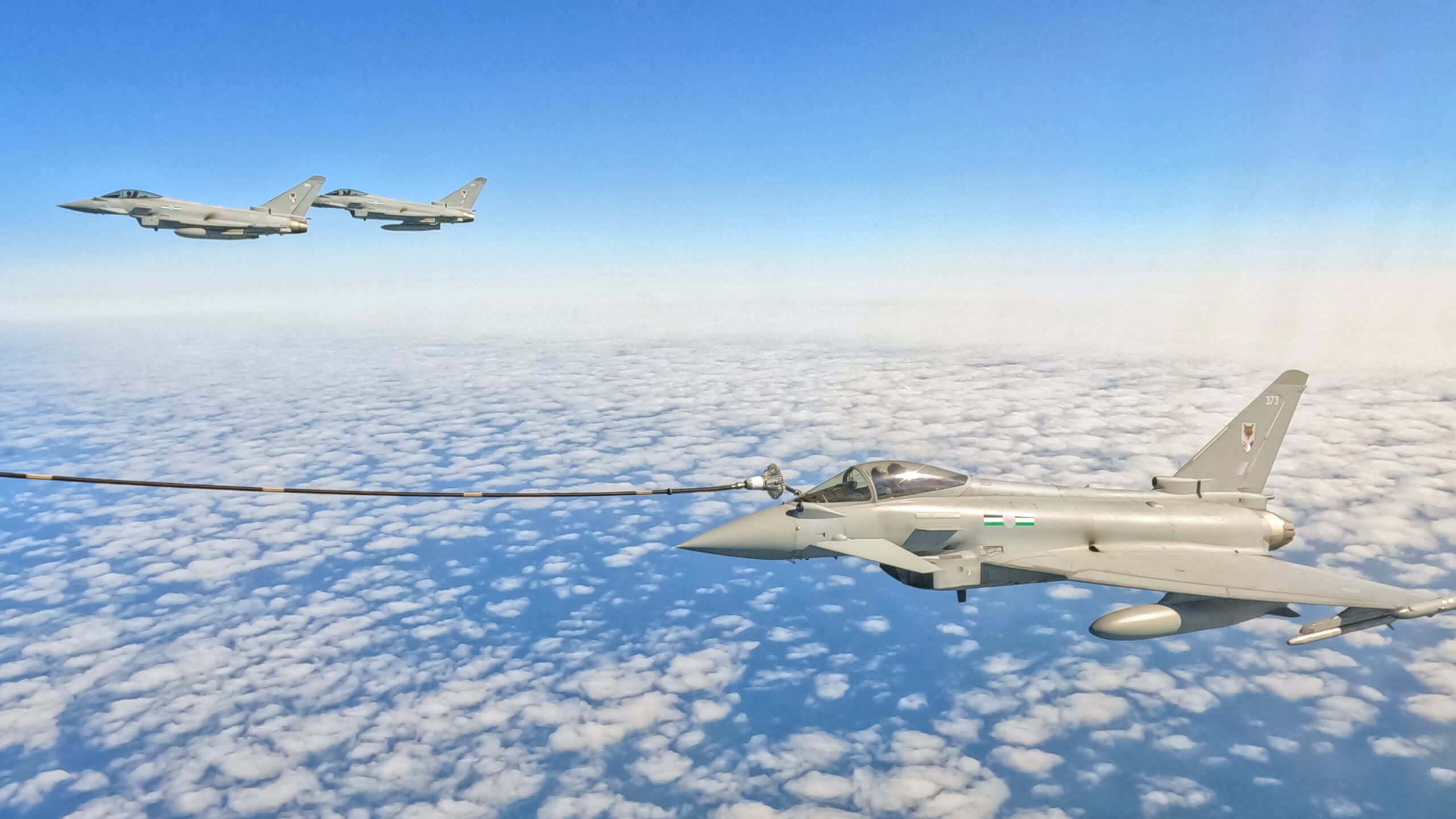 An RAF Typhoon fighter jet with a cable connected to it refuels in-flight over the North Sea. Two other jets can be seen in the background